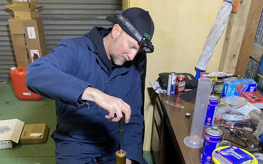 A man wearing blue coveralls, a black baseball hat and a headlamp sits nest to a table inside a repair garage turning a screwdriver on a piece of equipment in front of him.