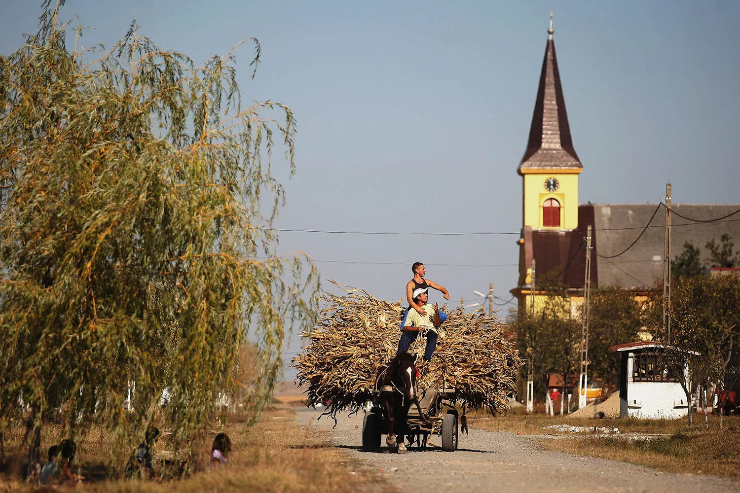 A long shot of two people riding on top of a horse-drawn cart stacked high with dried corn husks. They are traveling down a dirt road in a rural landscape, with a tall, yellow church tower featuring a clock visible in the background.