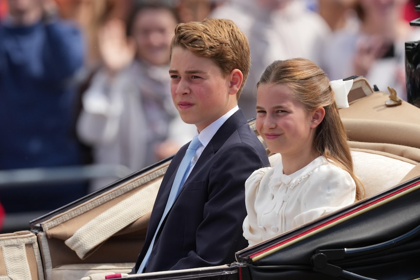 A boy and a girl in formal dress sit in a carriage as crowds look on.