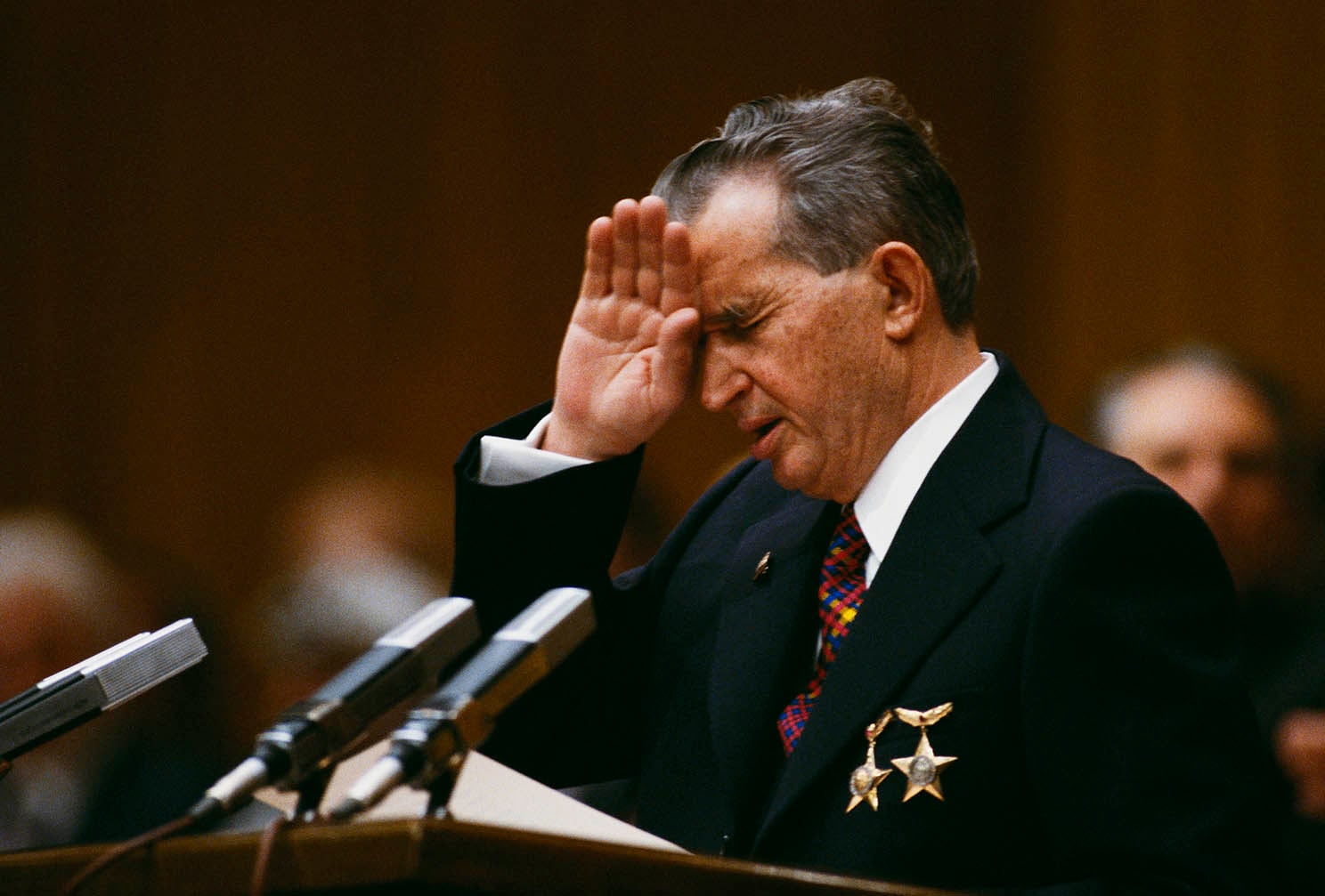 A man in a dark suit with medals pinned to his jacket, standing at a podium with microphones. He is holding his hand up near his forehead while speaking.
