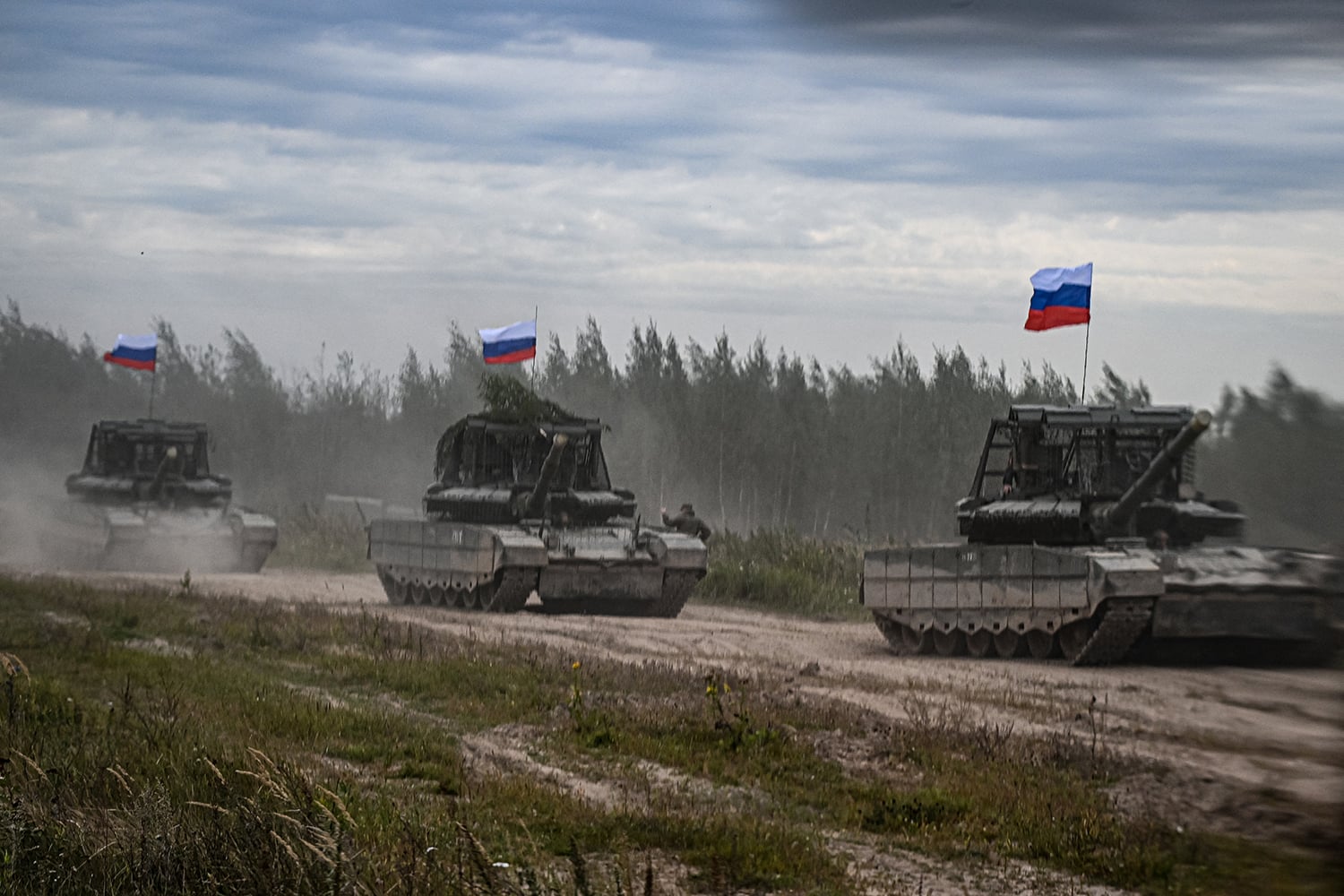 Three military tanks drive in a line along a dusty dirt road through an open field under a cloudy sky. Each tank flies a white, blue, and red horizontal tricolor flag from an antenna. A line of green trees is visible in the background.
