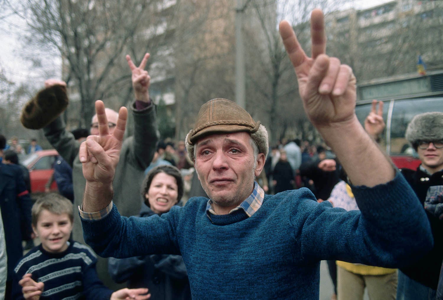 A crowd of people outdoors. In the center, a man in a blue sweater and a brown leather hat is looking forward with a tearful expression while raising both of his hands in V signs.