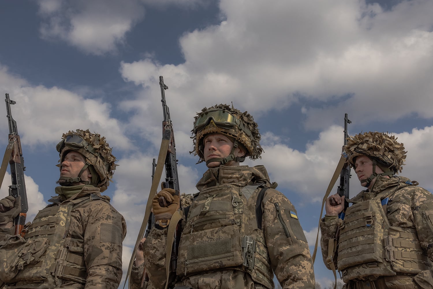 A low-angle shot of three soldiers standing outdoors against a blue sky with scattered clouds. The soldiers wear camouflage uniforms, helmets covered in textured foliage, and protective goggles. Each holds a rifle upright over their shoulder.