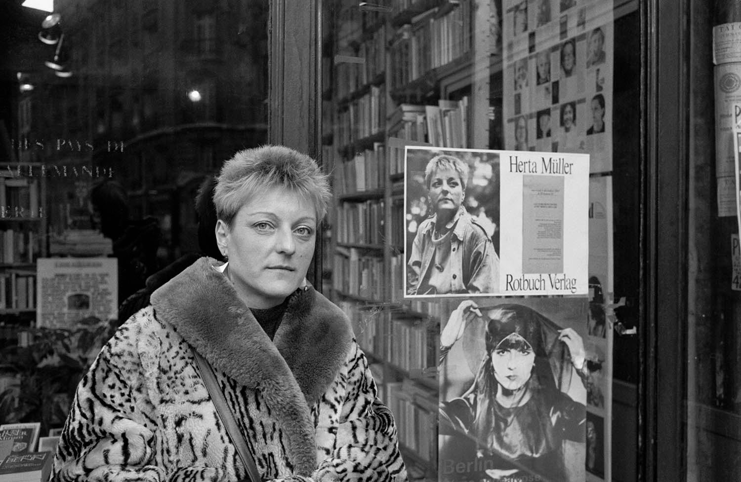 A black-and-white shot of a person with short hair wearing a leopard-print coat, standing in front of a bookstore window. Posters, including one featuring a portrait of the same person, are displayed on the glass.