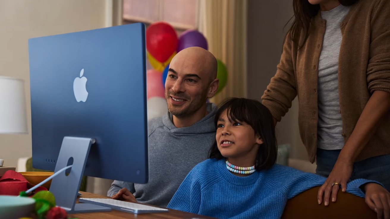 Bald man and child smiling while looking at a large blue Apple desktop computer, with a standing adult beside them and colorful balloons blurred in the background
