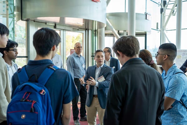 A white man in a blazer and khakis stands at the center of a group of students, speaking to them.