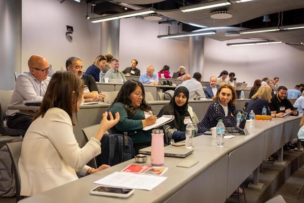 More students seated at long tables in the raked auditorium. Three women are in the foreground, one gesturing as she speaks, one writing on a tablet and one wearing a religious head covering.