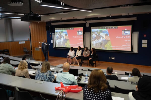 Students sit at tables in a raked auditorium, with four people seated onstage and a speaker at a podium. "Vision for AiBC" is projected on a screen.