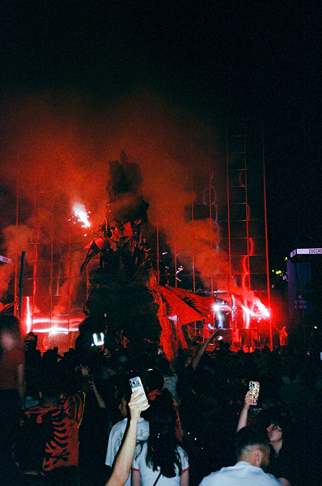 Crowd of protesters holding up cellphones under a large statue illuminated by red flares.