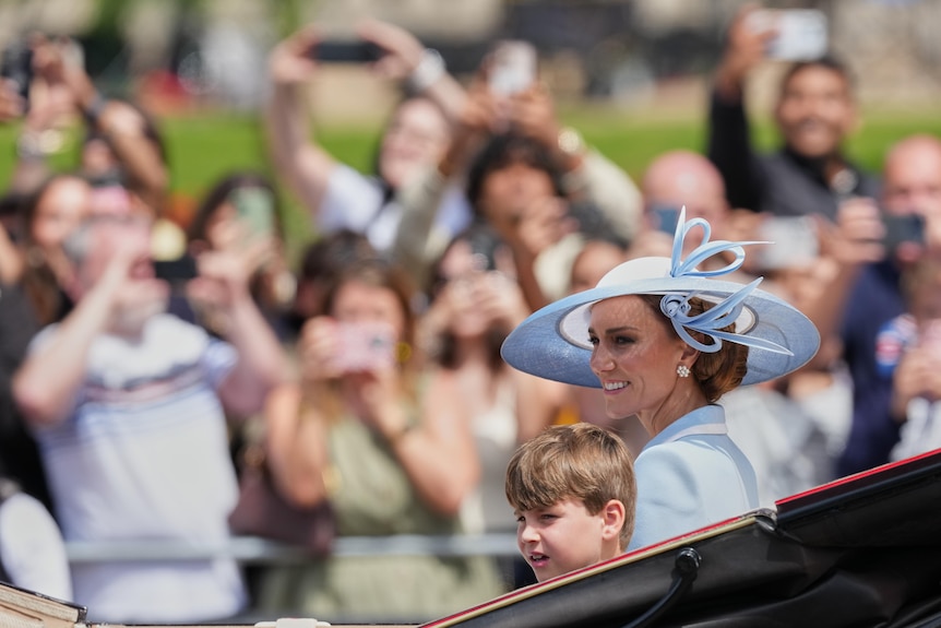Princess Catherine wearing a large sunhat and her son sit in a carriage while crowds look on taking photos.