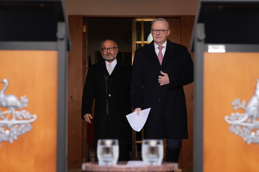 Two men in suits takl at a podium with an Australian flag behind them.