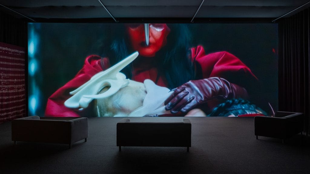 Large gallery projection displays a red-clad Björk beside sculptural mask forms in darkness.