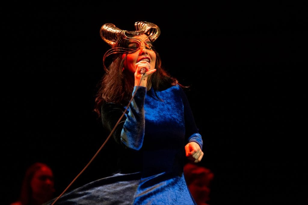 Björk performing onstage in a blue dress and elaborate gold mask by James Merry