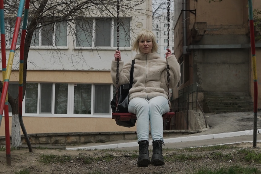 Tatiana sits on a swing with a straight expression.
