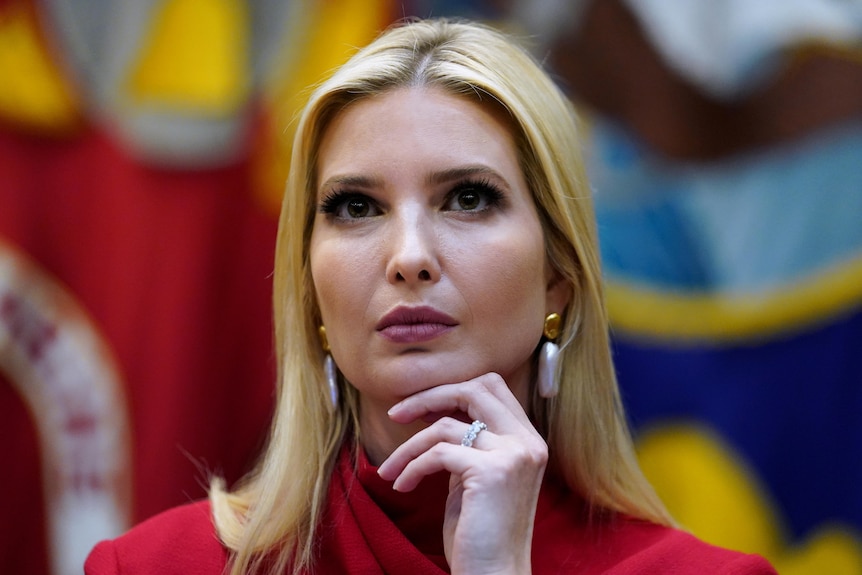 A close-up headshot of Ivanka Trump. She wears a red top and silver earrings. Her hand rests on her chin.