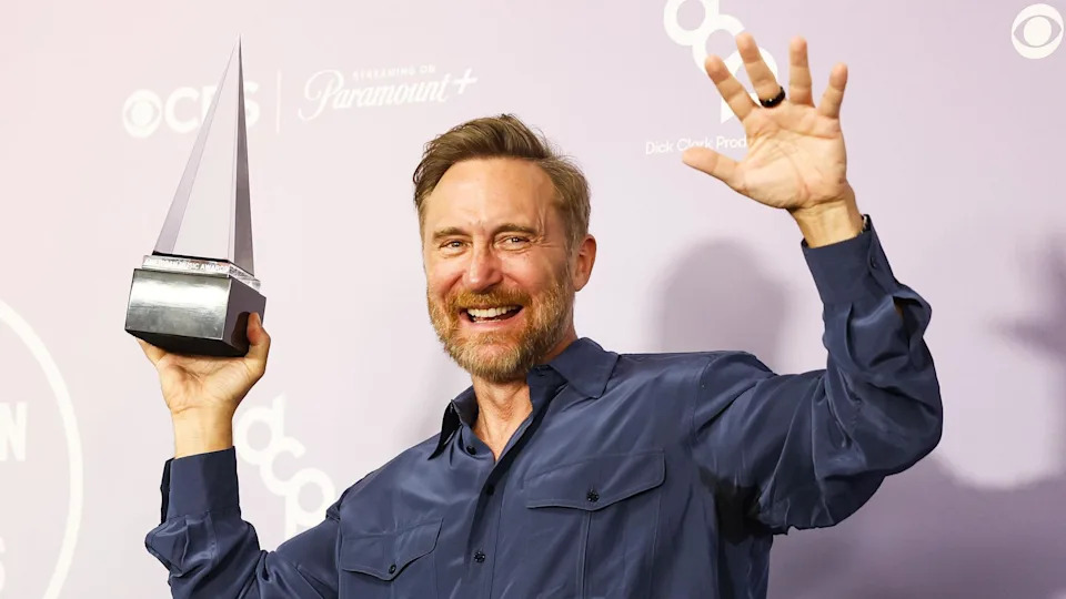 David Guetta holding an American Music Awards trophy which is triangular in shape. He is wearing a longsleeved blue buttoned shirt with pockets on the chest.
