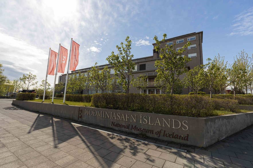 The exterior of the National Museum of Iceland (Þjóðminjasafn Íslands) in Reykjavik, with its sign and red institutional flags in the foreground.