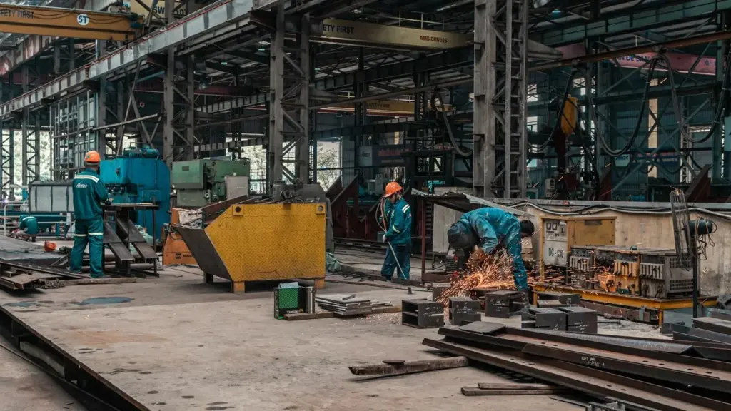 Darbininkai su apsauginiais šalmais dirba dideliame pramoniniame ceche, pjaudami metalo detales kampiniu šlifuokliu.