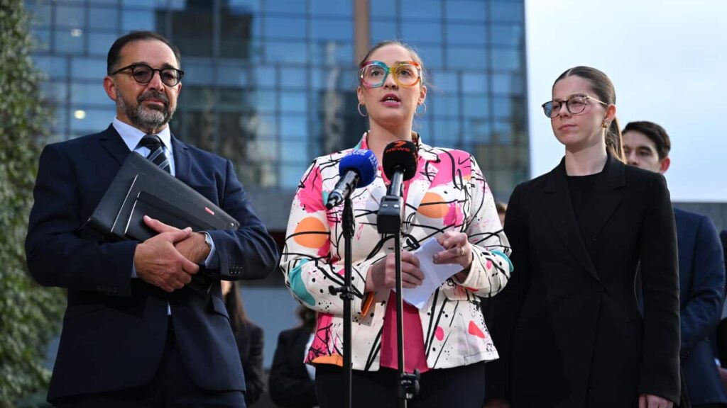 Robodebt victims share in millions as Australia’s largest-ever class action settlement approved Three people, a man and two women, stand before a pair of microphones outdoors. The man and woman on the left and right are dressed in dark suits while the woman in the centre is dressed in a colourful blazer and eyeglasses.
