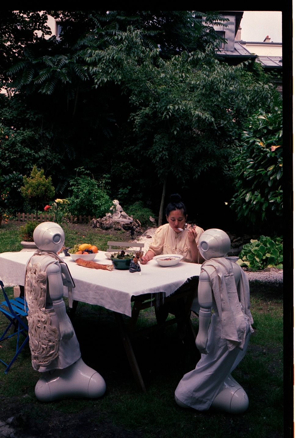 An outdoor scene shows a woman eating at a garden table with two small humanoid robots seated nearby, surrounded by dense greenery and domestic objects.