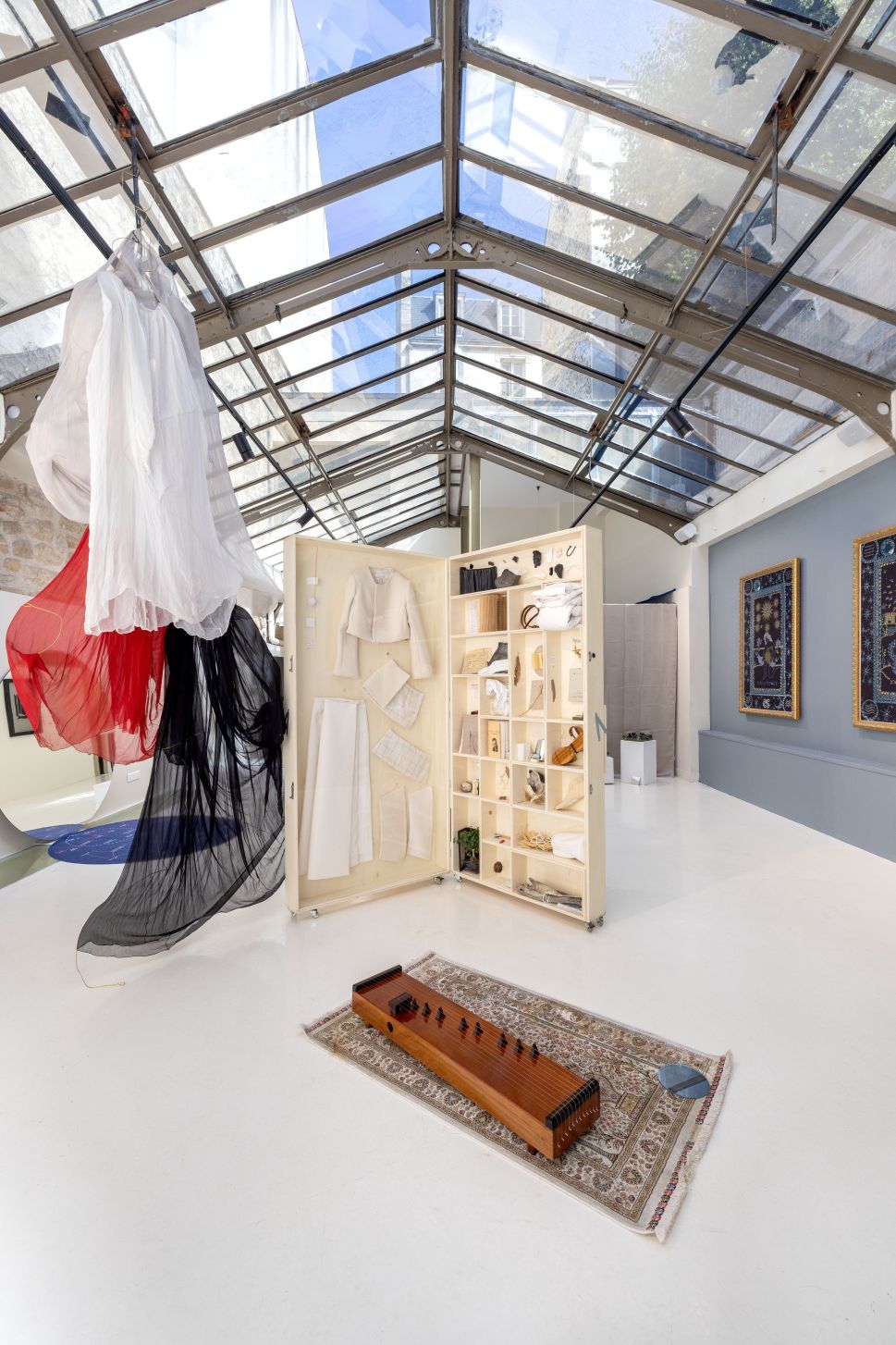 A bright gallery installation shows hanging translucent garments, an open wooden archive box filled with clothing and objects, a stringed instrument on a rug and framed textile works on the wall.