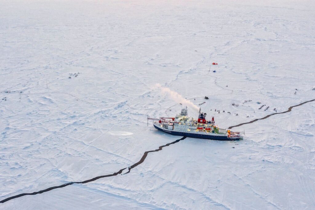 An icebreaker ship navigates through a cracked, frozen sea with scattered equipment and tents visible on the ice.
