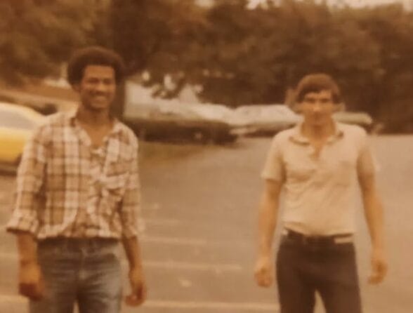 Two men walk toward the camera in a parking lot; one wears a plaid shirt and jeans, the other a polo shirt and jeans, with cars in the background (vintage photo).