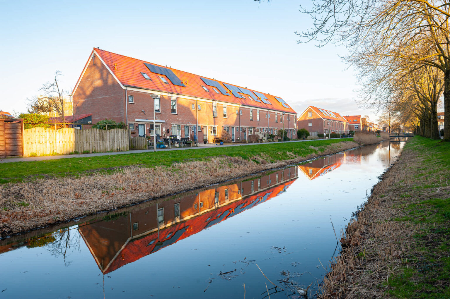 Netherlands solar owners face a harsh new math in 2027: Use midday power or lose money Residential buildings with red rooftops reflected in a calm canal, lined by trees and grassy banks.