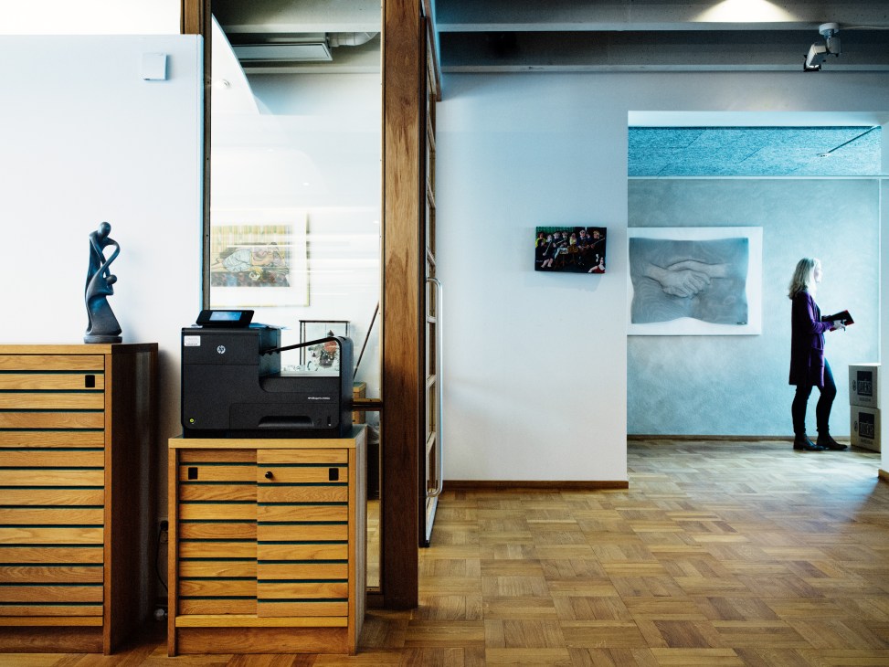 Woman viewing artwork displayed on white walls in a bright, modern office gallery space with wooden cabinets and a sculpture.