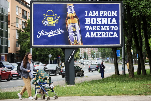 A woman walks past a billboard displaying lyrics from the Dubioza Kolektiv song "I Am From Bosnia, Take Me to America" in Sarajevo, Bosnia, Thursday, June 11, 2026. (AP Photo/Armin Durgut)