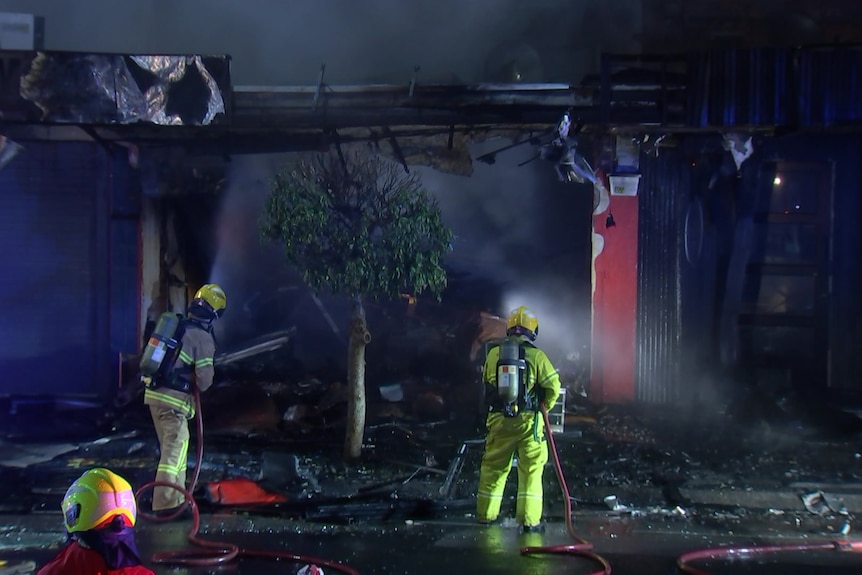 Two firefighters spray water into the burnt out shell of a store at night.