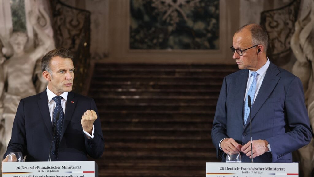 Americans Should Cheer Franco-German Nuclear Cooperation Two men in suits stand at podiums, speaking at a formal event. One gestures with a closed fist while the other looks at him. Ornate stairs and decor appear in the background.