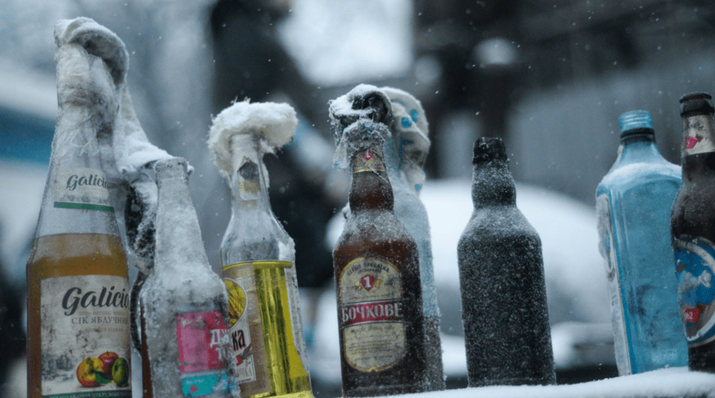 Several glass bottles covered in frost and snow are lined up outdoors in a cold, snowy environment. The bottles vary in color and label design, with some having caps wrapped in snow or ice. The background is blurred, emphasizing the frozen bottles in the foreground.