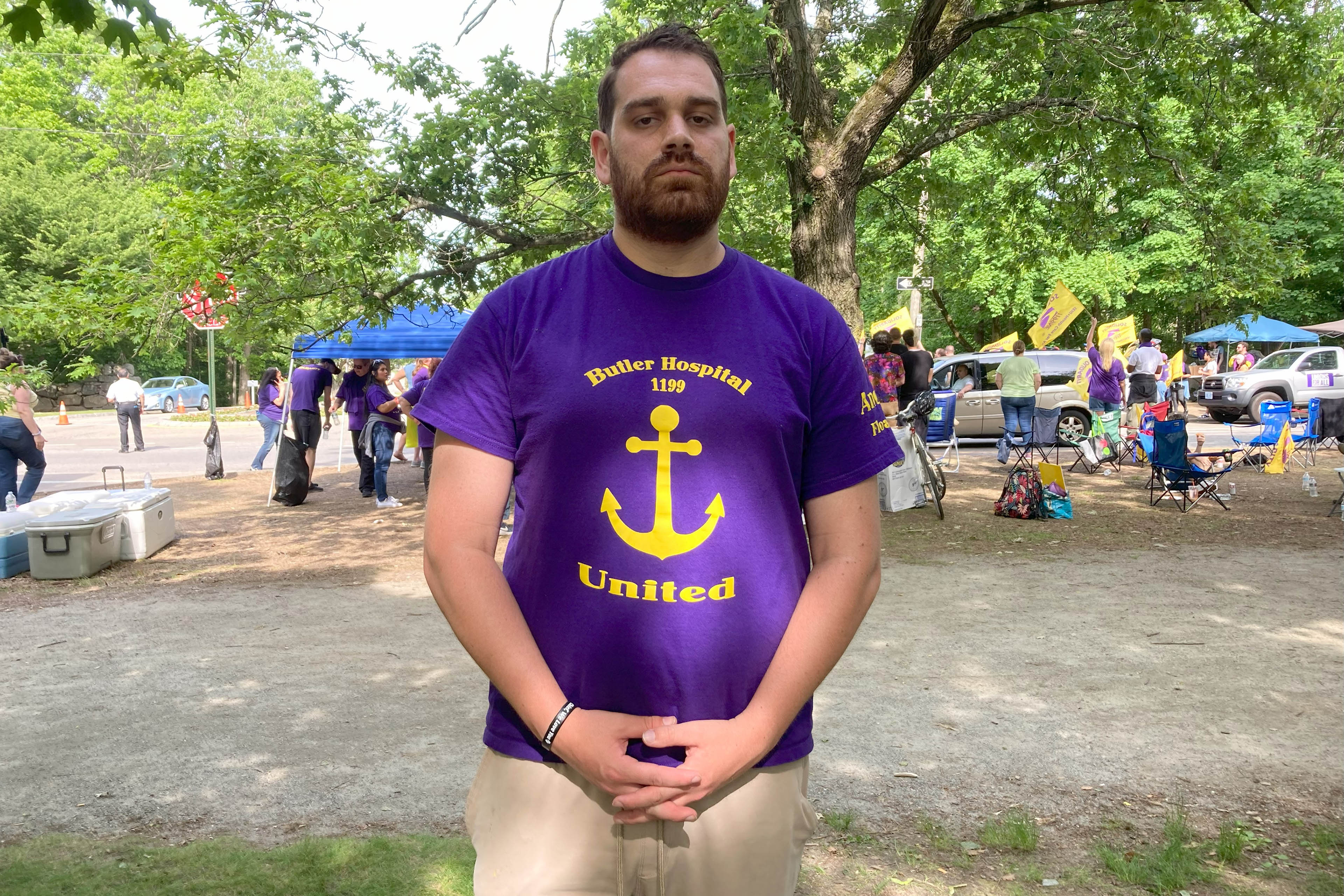 A man wearing a purple T-shirt that reads, "Butler Hospital 1199 United" stands for a photograph.