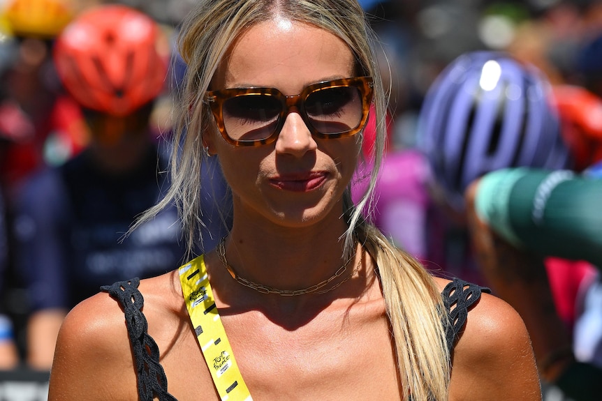 A blonde woman in sunglasses and a black top stands in the sunshine. 