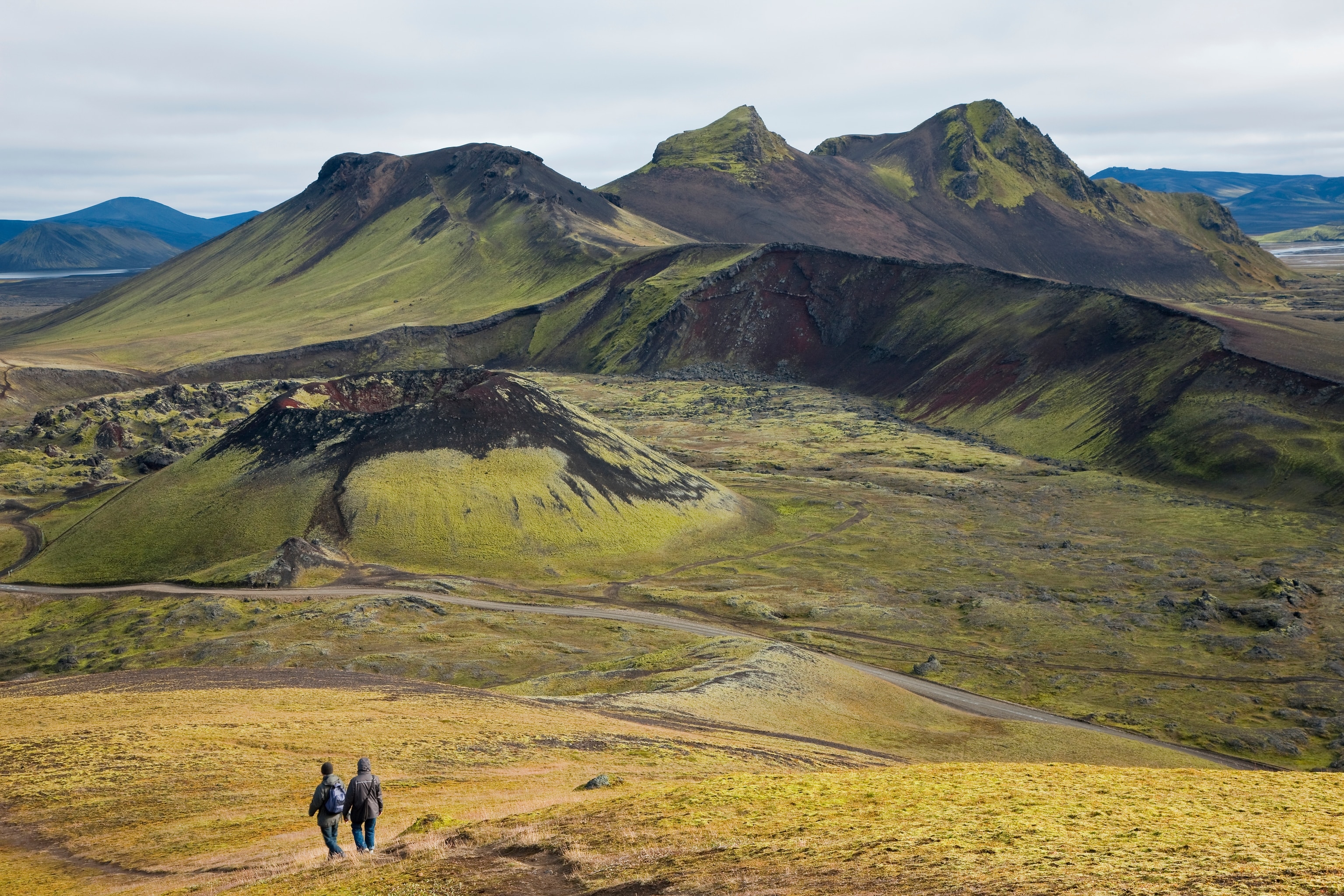 Two hikers walk towards large green mountains