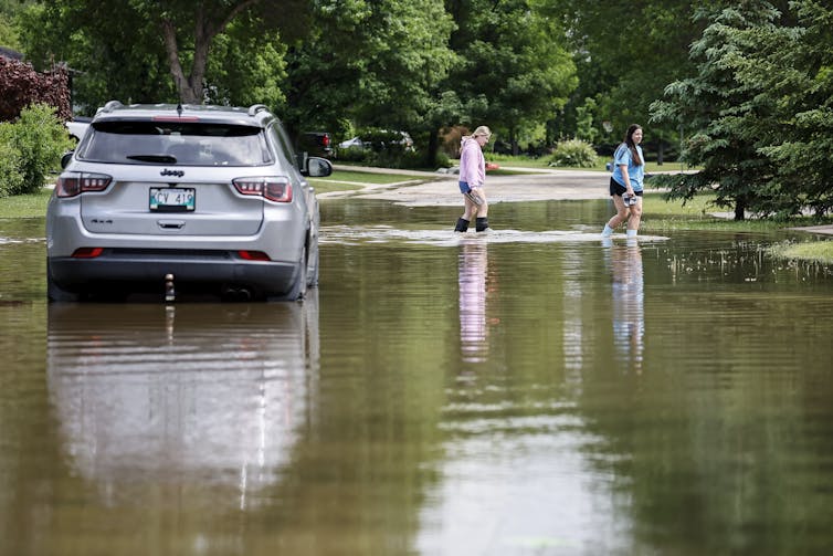 People walk by a flooded and abandoned vehicle on a street