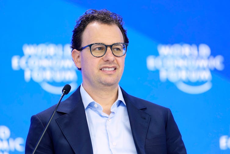 A white man with short, curly hair and glasses stands before a microphone