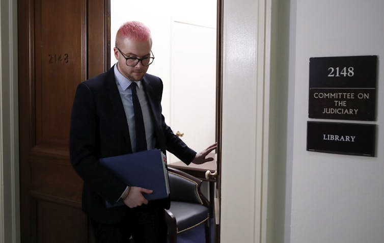 A white man with glasses and short, pink hair leaves a meeting room