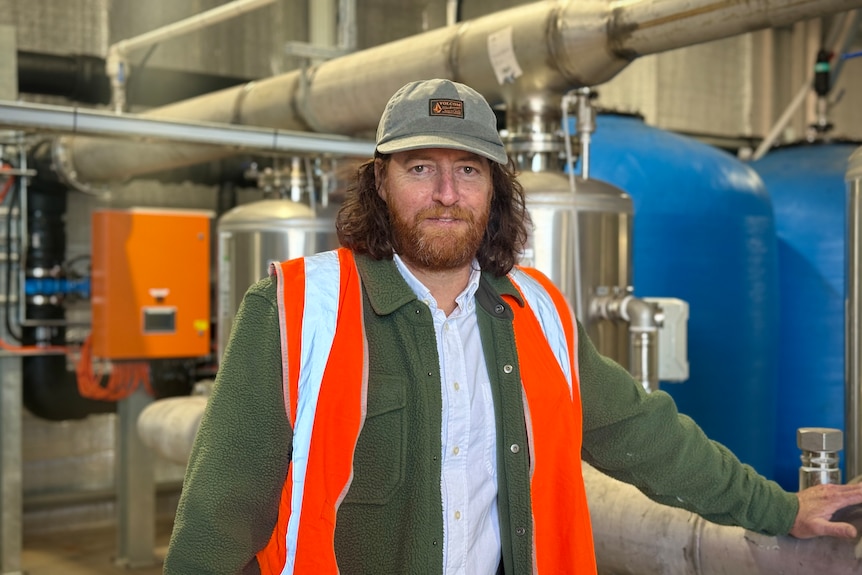 A middle-aged man with long hair and wearing a baseball cap stands in a water processing plant
