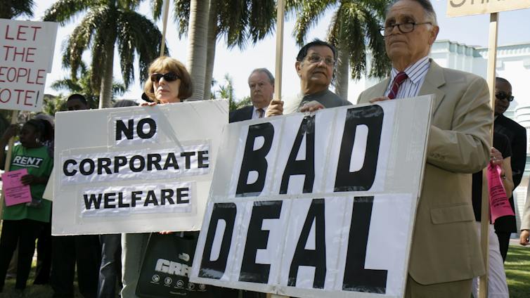 Protesters in Miami, with palm trees behind them, hold signs opposing the Marlins baseball stadium construction deal, citing taxpayer cost.