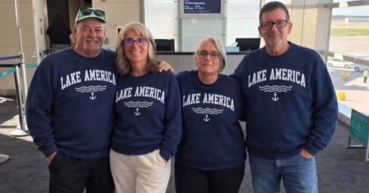 Four people stand side-by-side in an airport terminal wearing matching navy blue sweatshirts that say "LAKE AMERICA" with a wave and anchor graphic.