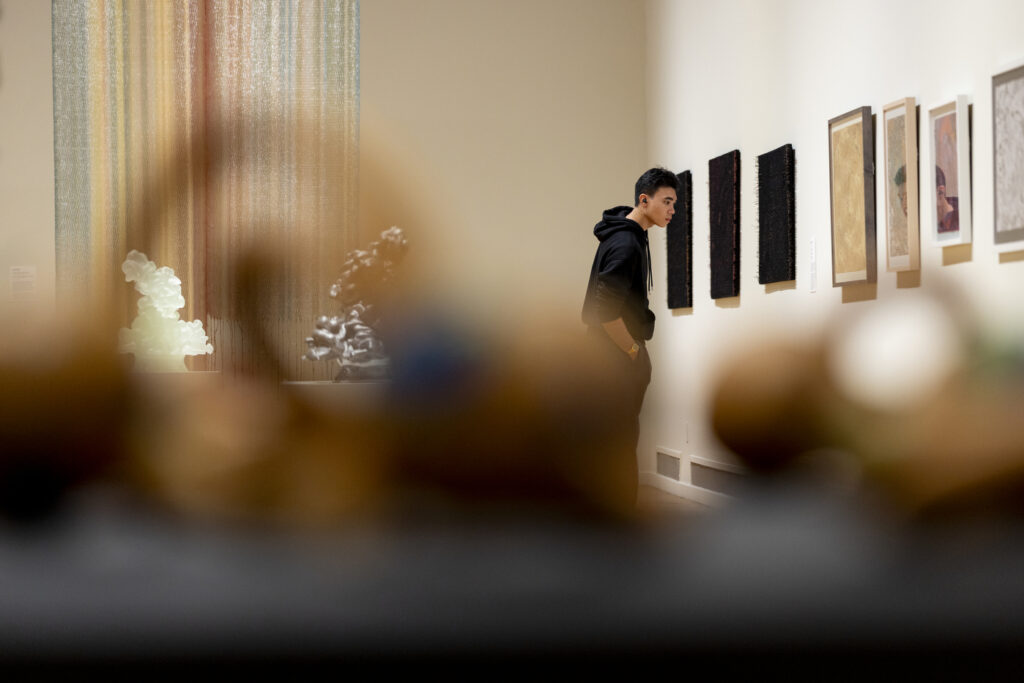 A student in a black hoodie studies a row of framed works on a gallery wall, seen past blurred objects in the foreground, with a tall multicolored hanging piece and small sculptures on a pedestal behind him.