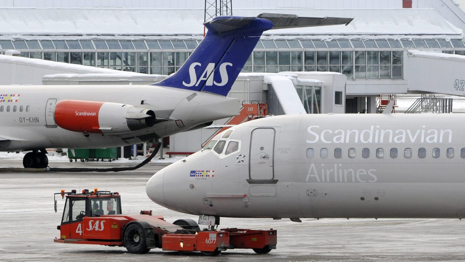 Two airplanes at an airport, one with a blue tail and the other labeled "Scandinavian Airlines," with a tug vehicle nearby.