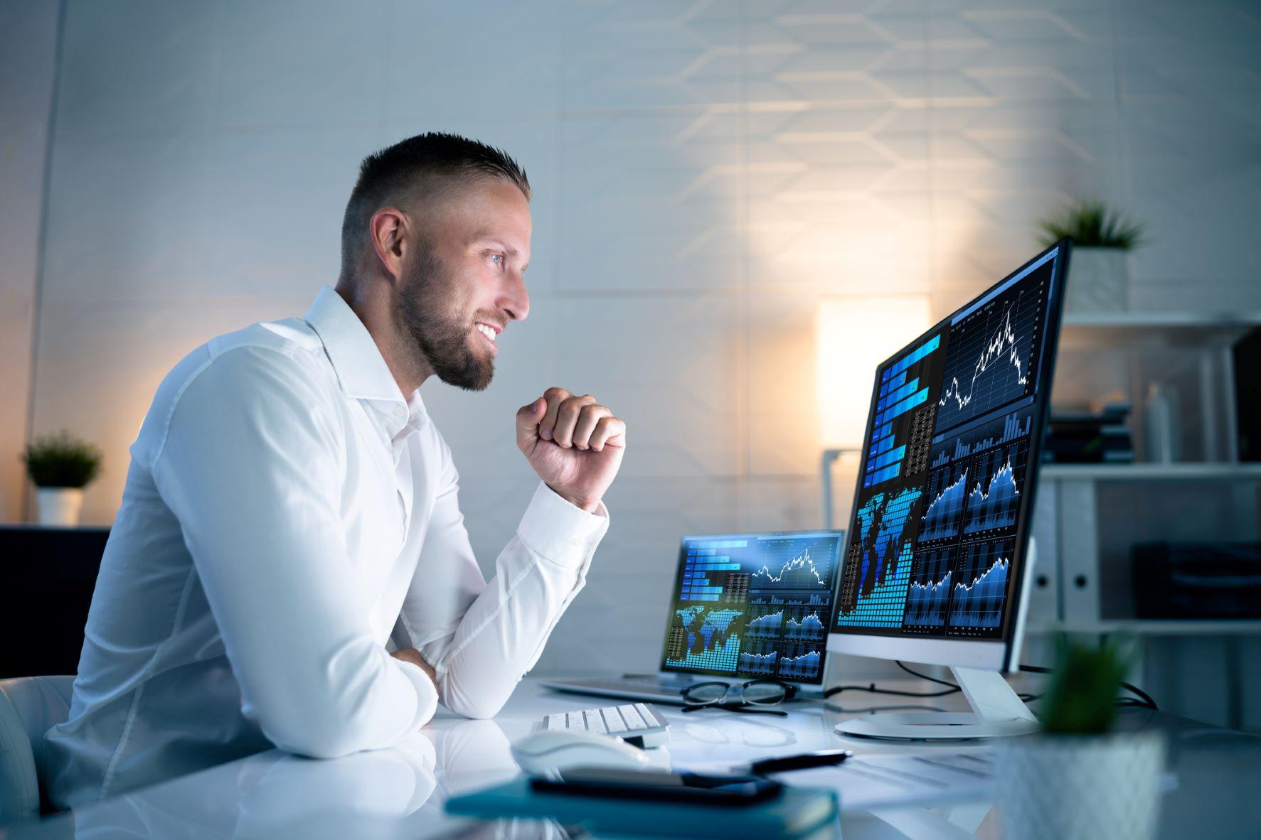 Smiling financial analyst monitors stock market charts on multiple screens in a modern office.