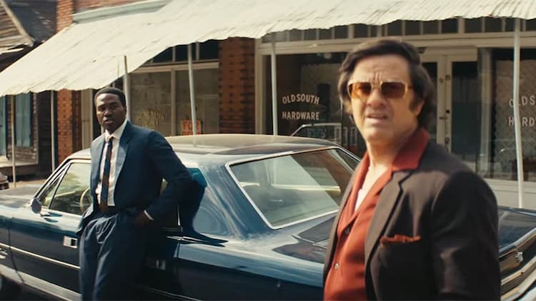 Strider and Scarpa are on the street. Strider is leaning against his car. Scarpa is looking around the area. They are outside a row of shops.