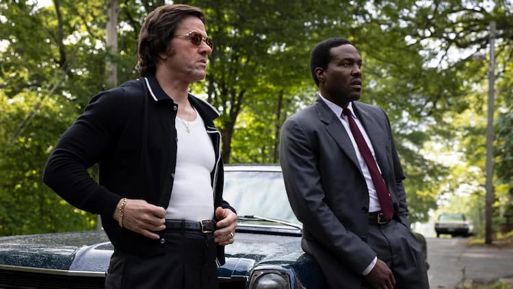 Scarpa and Strider are next to a car in the suburbs. Strider is leaning against the car with one hand in his pocket. Scarpa is next to the car, fixing his jacket. Trees are in the background.
